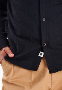 Niebieska koszula w jodełkę wykonana z miękkiego materiału, z brązowymi guzikami i metką boczną z logo, w połączeniu z beżowymi spodniami.