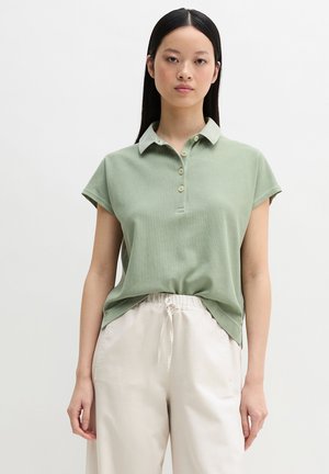 Junge Frau, die ein hellgrünes Polohemd mit kurzen Ärmeln und cremefarbene Hosen mit Kordelzug trägt, steht vor einem schlichten, hellen Hintergrund.