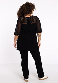 Schwarze Tunika mit transparenten Glockenärmeln, die am Rücken mit gestreiften Details versehen ist. Kombiniert mit schwarzen Hosen und Pantoletten mit einer beigen Sohle.