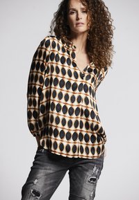 Musterbluse in Creme mit schwarzen ovalen Formen, die in Orange umrandet sind, mit V-Ausschnitt und Ballonärmeln, kombiniert mit ausgewaschenen schwarzen Jeans.