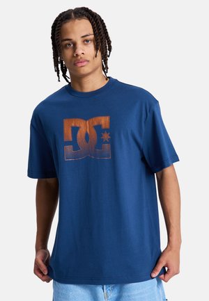 Junger Mann mit geflochtenem Haar, der ein blaues T-Shirt mit einem großen braunen DC-Logo trägt und vor einem schlichten weißen Hintergrund steht.