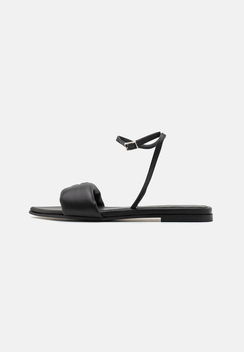 Svart platt sandal med vadderad framrem, tunn ankelsrem med silverfärgad spänne och fyrkantig tå-sula på vit bakgrund.