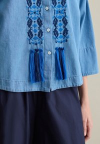 Camicia di denim blu chiaro con intricati ricami blu scuro e frange blu, dotata di chiusura a bottoni e maniche a tre quarti.