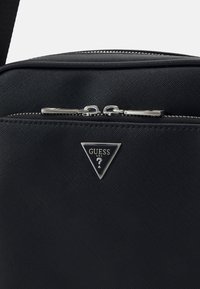 Schwarze Nylon-Crossbody-Tasche mit einem Reißverschluss, ausgestattet mit einem dreieckigen Metall-Logo-Schild und silbernen Hardware-Akzenten. Strukturierte Oberfläche.