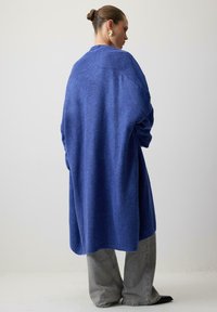 Naine, kelle juuksed on pundis, kannab suuruses suuri sinist kudumisseelikut, halli laiade pükse ja kuldseid geomeetrilisi kõrvarõngaid, seistes lihtsa valge tausta ees.