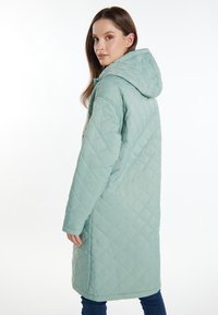 Giacca lunga trapuntata verde chiaro con cappuccio, caratterizzata da una texture liscia e un motivo a diamante, mostrata di spalle con un profilo laterale.