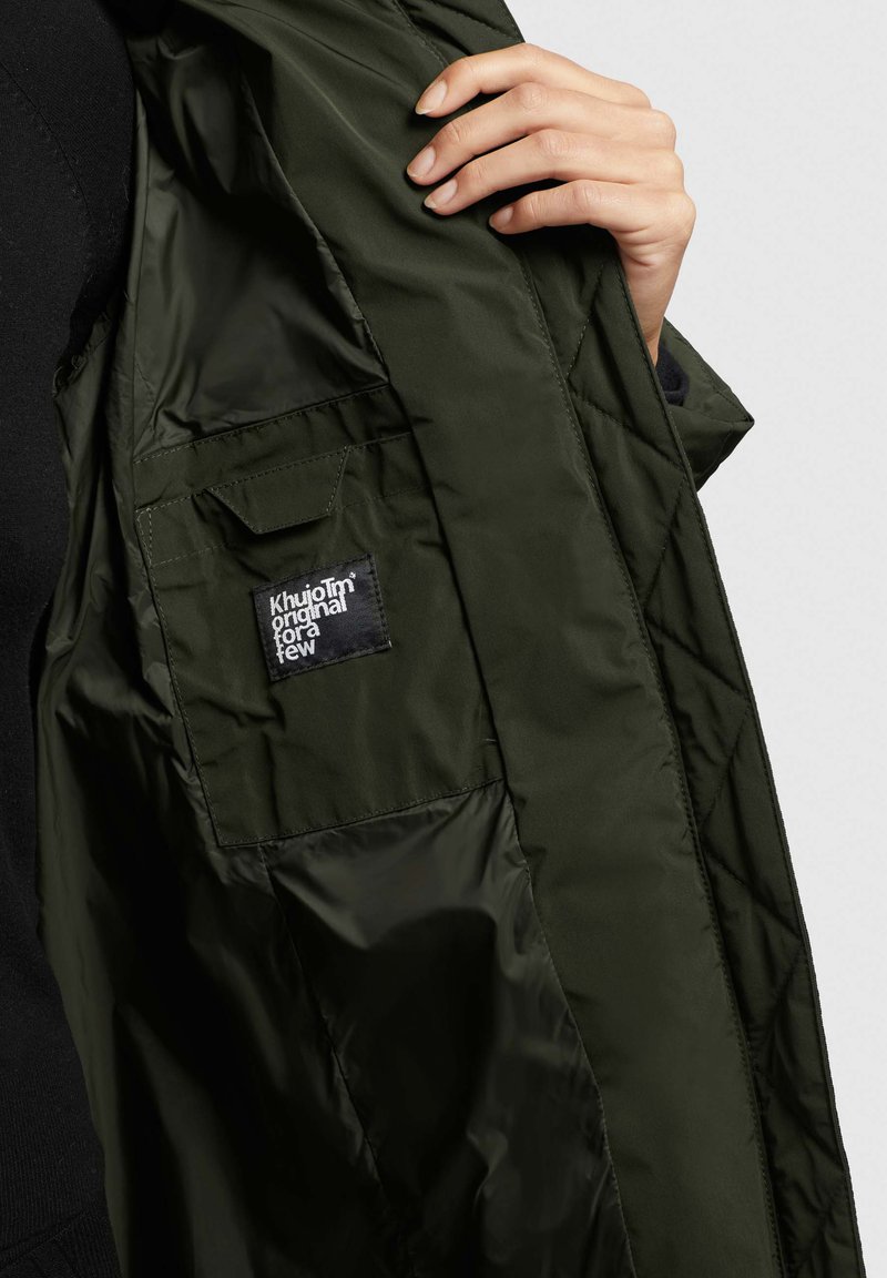 Dunkelgrüne gesteppte Jacke mit einer Tasche und einem schwarzen Etikett. Das Innere ist glänzend mit einer glatten Textur. Nahaufnahme des inneren Designs.