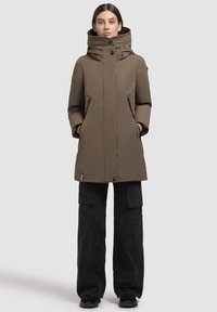 Femme portant un manteau d'hiver à capuche marron avec des poches zippées noires et un pantalon cargo noir, se tenant avec les mains dans les poches sur fond blanc.