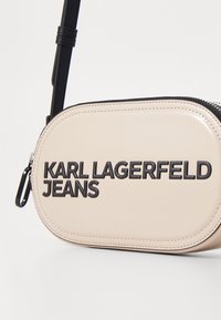 Borsa ovale in pelle beige con zip nera, logo nero "KARL LAGERFELD JEANS" e tracolla regolabile. Texture liscia con dettagli cuciti.