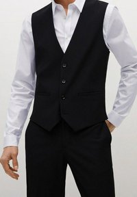 Gilet nero su misura con un design aderente, dotato di quattro bottoni, due tasche frontali e una superficie liscia, indossato sopra una camicia bianca.