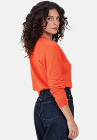 Orangefarbener Strickpullover mit gerippten Bündchen und Saum, kombiniert mit dunkelblauen Jeans aus Denim. Der Pullover hat eine lockere Passform und einen Rundhalsausschnitt.