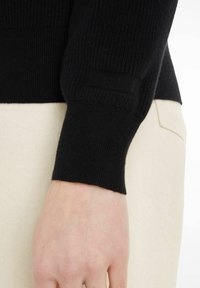 Close-up af en persons håndled iført en sort ribbet sweaterærme over beige bukser.