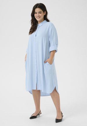 Vestido camisero azul claro con botón en la parte delantera, mangas enrollables, bolsillos laterales y un dobladillo curvado. Fabricado con una tela suave.
