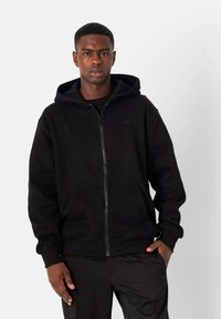 Sudadera con cremallera negra hecha de una mezcla de algodón. Presenta un bolsillo tipo canguro y una capucha forrada. Sutil logotipo tonal en el pecho.