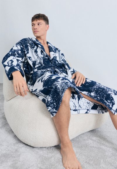 Een marineblauwe en witte badjas met een capuchon en ceintuur, gemaakt van zachte terrycloth, gedragen door een persoon die ontspant op een pluche zitzak.