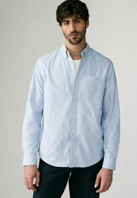 Chemise à boutons bleu clair et blanche à rayures verticales, à manches longues, avec un col retroussé et une poche poitrine. Le tissu semble lisse.