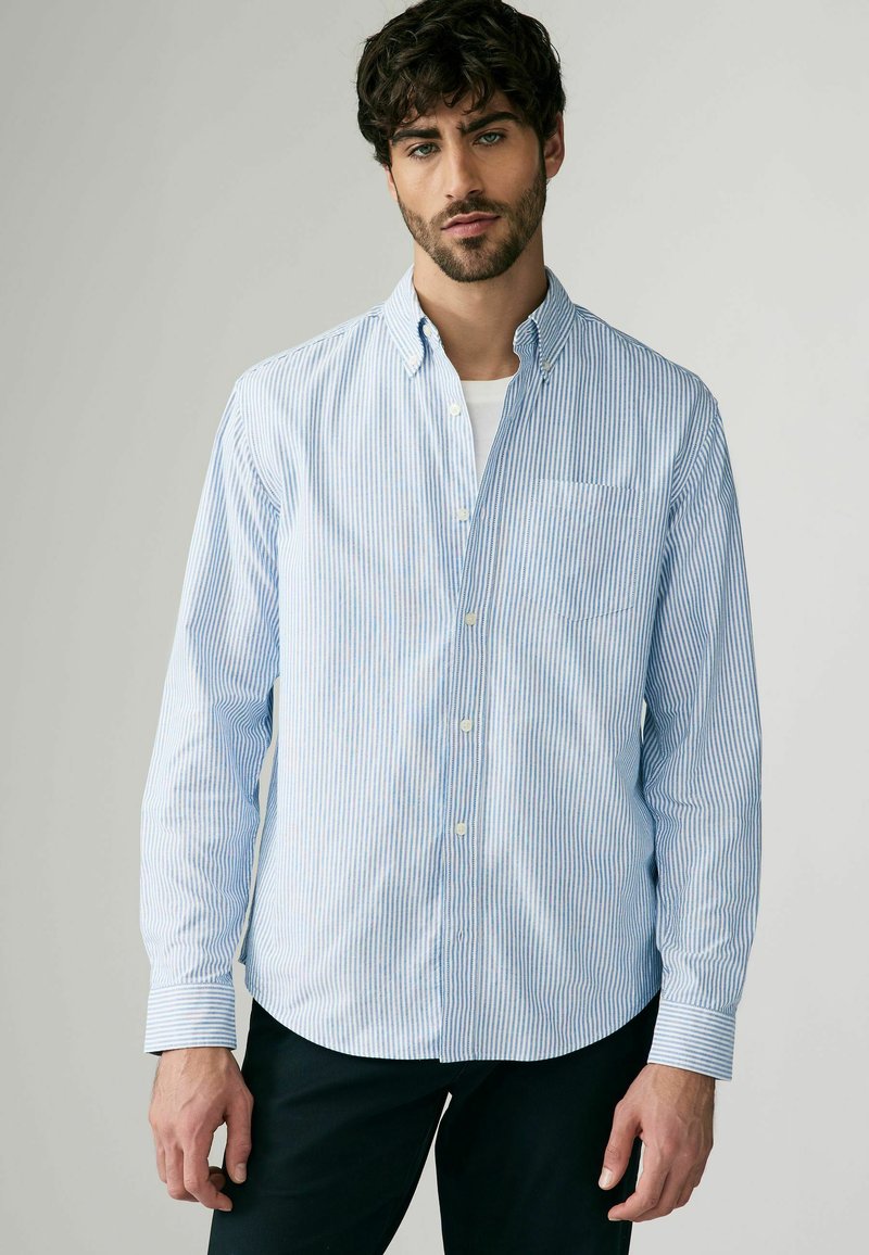 Chemise à boutons bleu clair et blanche à rayures verticales, à manches longues, avec un col retroussé et une poche poitrine. Le tissu semble lisse.