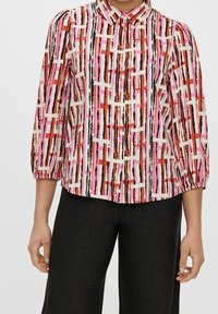 Blouse col classique avec des manches bouffantes, ornée d'un motif géométrique multicolore dans des nuances de rose, rouge, noir et blanc. Design à boutons sur le devant.