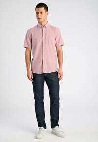 Kortärmad, ljusrosa knäppskjorta i ett texturerat tyg, kombinerad med mörkblå tajta jeans och vita sneakers.
