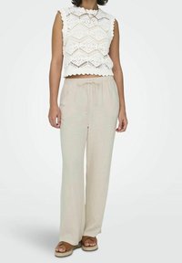Haut blanc court avec un motif en dentelle hexagonal et des bords festonnés, associé à un pantalon large beige léger et des sandales marron.