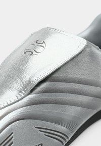 Ezüst bebújós cipő texturált felsőrésszel, amely vízszintes vonalakat és márkás díszítést tartalmaz. A kialakítás tépőzáras pántot is magában foglal.