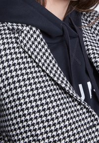 Blazer de pata de gallo en blanco y negro llevado sobre una sudadera negra con cordones, mostrando parcialmente el pecho y el cuello de una persona.