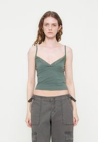 Femme aux cheveux ondulés portant un caraco vert bordé de dentelle et un pantalon cargo gris, debout devant un fond blanc uni.