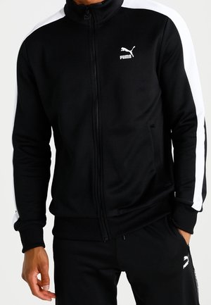 Mężczyzna w czarnym dresie Puma z białymi paskami na rękawach i spodniach, stojący z jedną ręką w kieszeni.