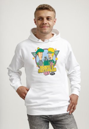 Mann trägt weißen Hoodie mit buntem Beavis & Butt-Head '93 Motiv, Hände in der vorderen Tasche, schaut seitlich mit leichtem Lächeln.