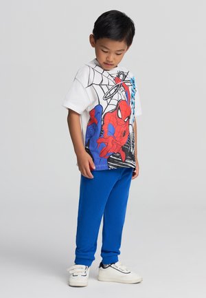 T-shirt bianco in cotone con un design grafico di Spider-Man colorato, abbinato a pantaloni tuta blu in cotone, indossato con sneakers bianche.