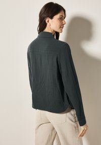 Camicia a maniche lunghe di un verde scuro, con una texture, colletto e polsini abbottonati, abbinata a pantaloni beige chiaro.