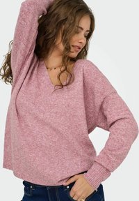 Jeune femme aux cheveux bouclés portant un pull en tricot rose et un jean bleu, posant avec une main sur la hanche et l'autre levée.