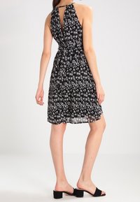 Black sleeveless dress with white floral pattern, cinched waist, open back, and knee-length skirt. Worn with black block-heeled sandals.
