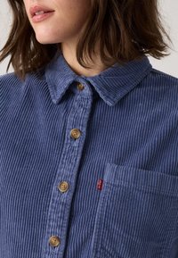 Blauwe corduroy shirt met een spitse kraag, zeven plastic knopen, borstzak en subtiele ribbelstructuur. Rode logotag op de zak.