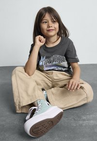 Bambino con una maglietta nera con grafica che presenta "Converse", pantaloni larghi di velluto a coste beige e scarpe alte verde menta con suole bianche.
