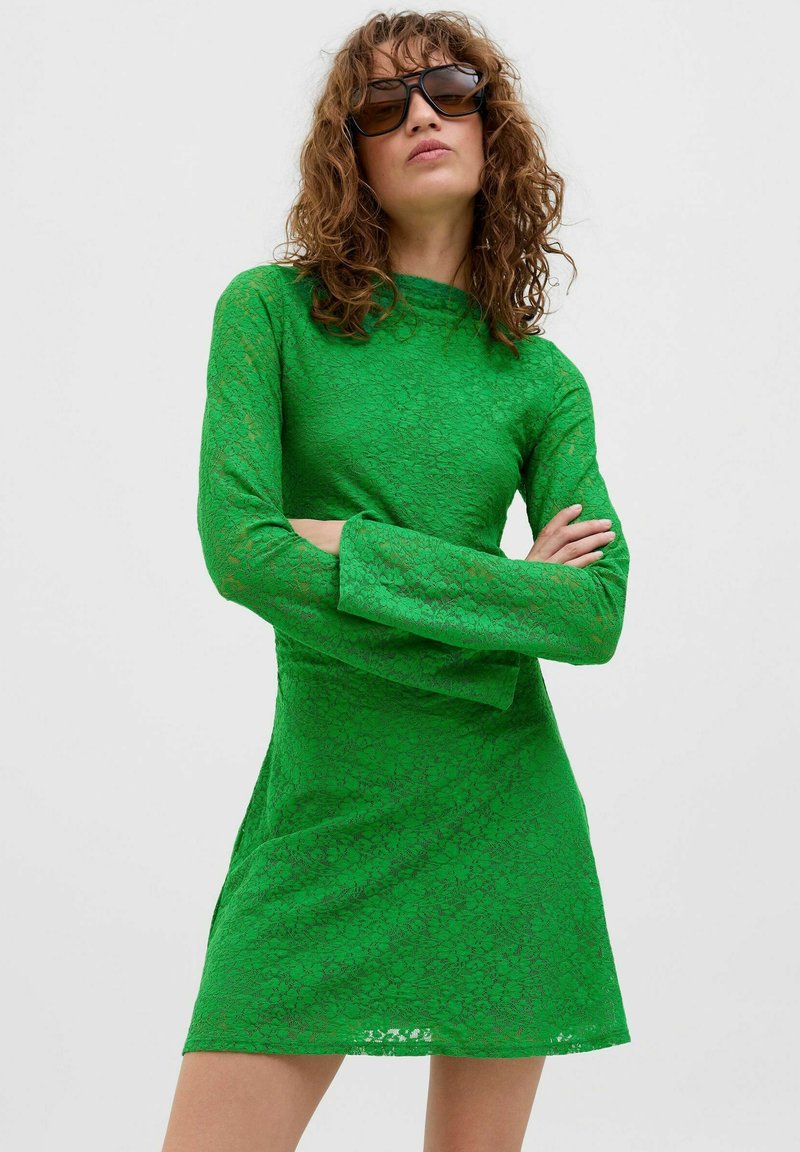 Robe mini à manches longues en dentelle verte avec un col montant, silhouette ajustée et motif floral. Le mannequin porte des lunettes de soleil.