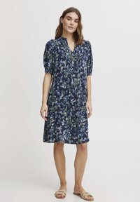 Mornarska cvetna obleka s napihnjenimi rokavi, V-izrezom in dolžino do kolen. Ima zelene in bele cvetlične vzorce na temni podlagi.