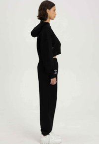 Zwarte cropped hoodie met een capuchon, gecombineerd met hoge zwarte joggingbroek. Beide hebben een gladde textuur; de outfit heeft minimale branding.