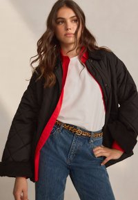 Black quilted jacket with red accents, white shirt, and blue denim jeans. Leopard print belt adds a distinctive pattern.