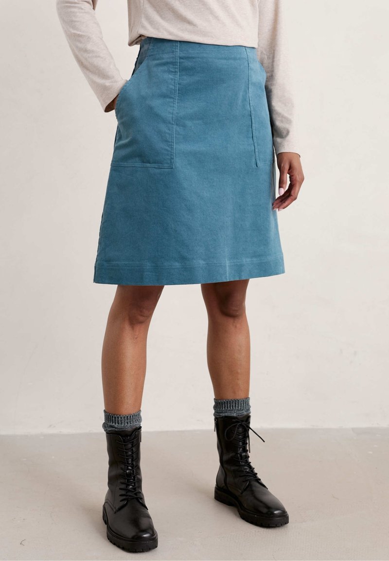 Personne portant une jupe bleue mi-longue avec de grandes poches avant, des chaussettes grises, des bottes noires à lacets et un haut beige clair à manches longues.