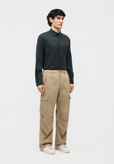 Άνδρας που στέκεται, φορώντας σκούρο πράσινο πουκάμισο με μακριά μανίκια και κουμπιά, μπεζ παντελόνι cargo και λευκά αθλητικά παπούτσια, μπροστά σε απλό φόντο.