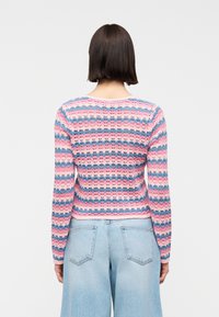 Γυναίκα με κοντά σκούρα μαλλιά που φοράει ζιβάγκο πουλόβερ με ριγέ μοτίβο σε ροζ και μπλε χρώματα και ανοιχτόχρωμο τζιν ψηλόμεσο, με την πλάτη προς τον θεατή.