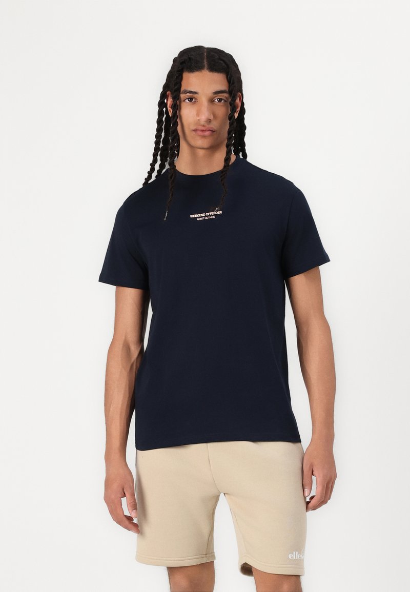 Mørkeblå bomulls T-skjorte med korte ermer, med en liten hvit tekstlogo på brystet. Kombinert med beige shorts.