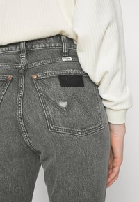 Jeans Wrangler z šedého denimu s vysokým pasem, které mají měděné nýty a černou koženou nášivku na zadní straně. Měkká struktura s mírným vyblednutím.