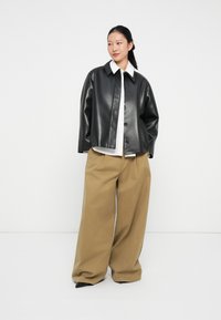 Giubbotto in ecopelle nera con design a bottoni, abbinato a pantaloni larghi beige a gamba larga. Il modello è in piedi contro uno sfondo bianco uniforme.