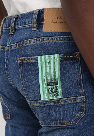 Jean en denim avec un lavage bleu et des coutures orange. Comprend une étiquette en cuir portant la mention "PS Paul Smith" et une étiquette d'entretien verte avec un code QR.