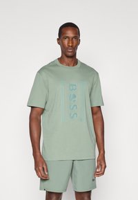 Tricoul din bumbac de culoare verde mentă are un logo tonal "BOSS" în centru. Modelul poartă şorturi asortate, tot de culoare verde mentă, ambele având o textură netedă.