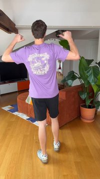 Man standing indoors wearing a purple "The Going Merry" shirt, black shorts, and white sneakers, pointing thumbs at the shirt's back.