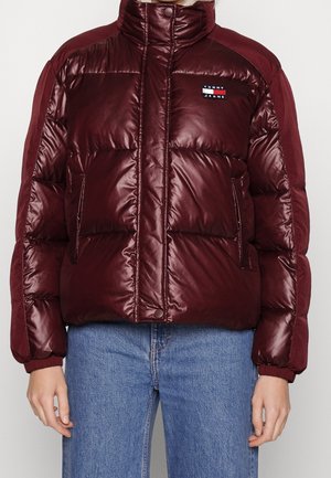 Bordeauxrote, gesteppte Steppjacke mit hohem Kragen, elastischen Bündchen, Seitentaschen und Tommy Jeans-Logo, getragen über blauen Denim-Jeans.