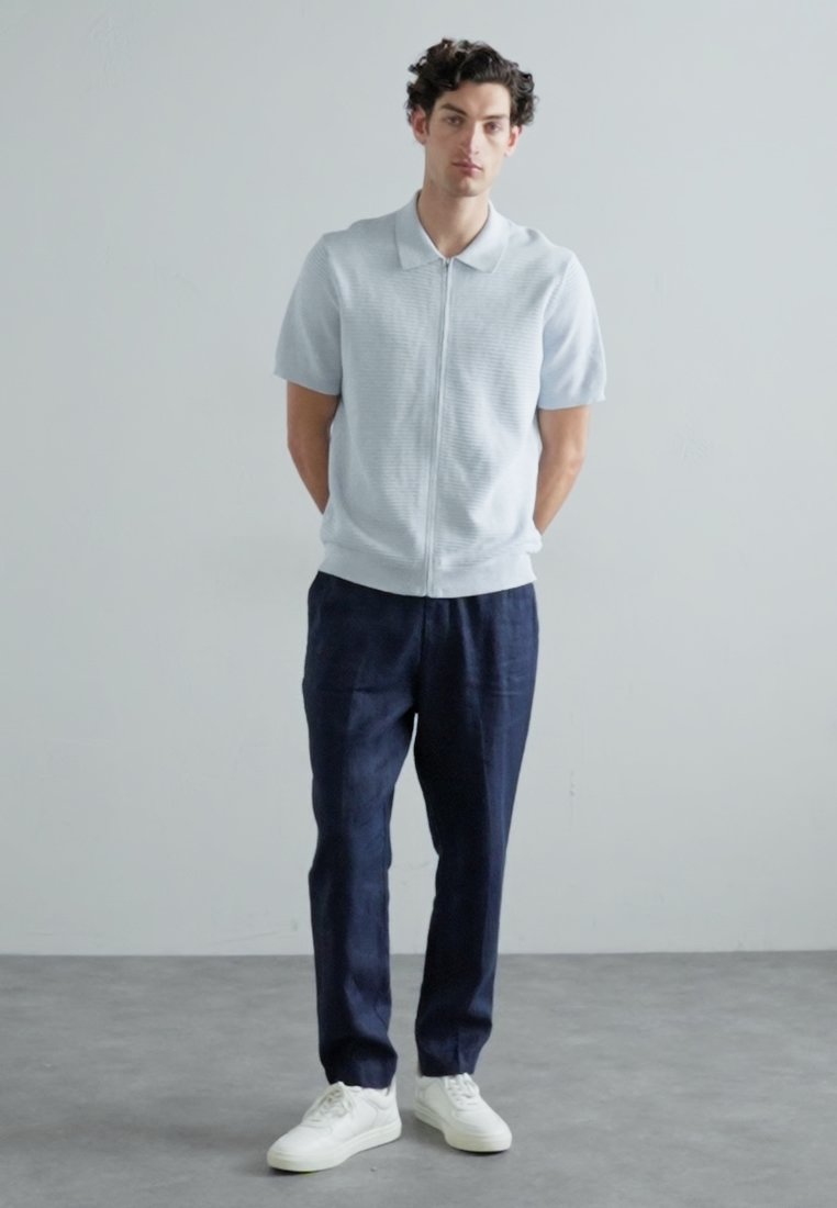 Polo bleu clair à manches courtes avec une fermeture éclair à l'avant, associé à un pantalon marine et des baskets blanches. Texture lisse, design simple et coupe décontractée.